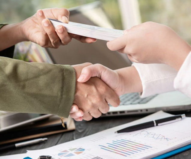 Hands exchanging documents with a handshake, symbolizing financial agreement and quick cash loans, with a laptop and charts in the background.