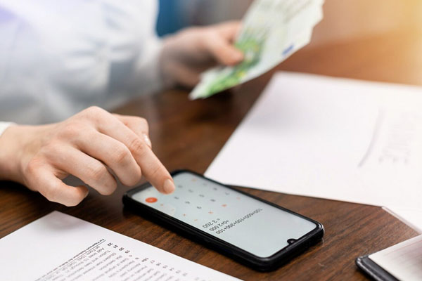 Person using smartphone to manage finances, holding cash, with documents on a table, representing instant funding payday loans.